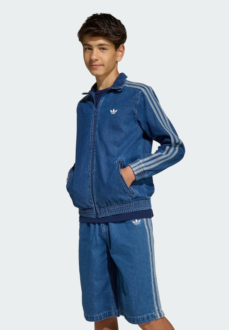 Completo tuta in denim composto da giacca con zip, due tasche frontali e strisce grigie, abbinata a pantaloncini coordinati. Colore blu su tutta la superficie.