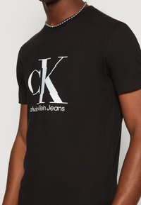 Černé bavlněné tričko s výrazným bílým logem Calvin Klein, kulatým výstřihem a krátkými rukávy. Viditelný texturovaný detail látky.