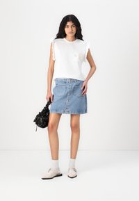 T-shirt en coton blanc avec des manches volantées et une poche avant ; mini-jupe en denim bleu avec des boutons ; sac à motifs noir ; chaussures beiges avec des lanières.