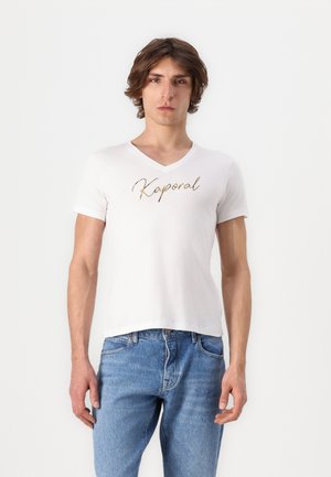 Jonge man met halflang haar, gekleed in een wit V-hals T-shirt met het opschrift "Kaporal" en een blauwe spijkerbroek, staande tegen een effen witte achtergrond.