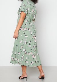 Eine Frau trägt ein grünes, floral gemustertes Midikleid mit kurzen Ärmeln und einer Schleife im Rücken, kombiniert mit schwarzen, offenen Absatzschuhen, und steht auf einem grauen Boden.