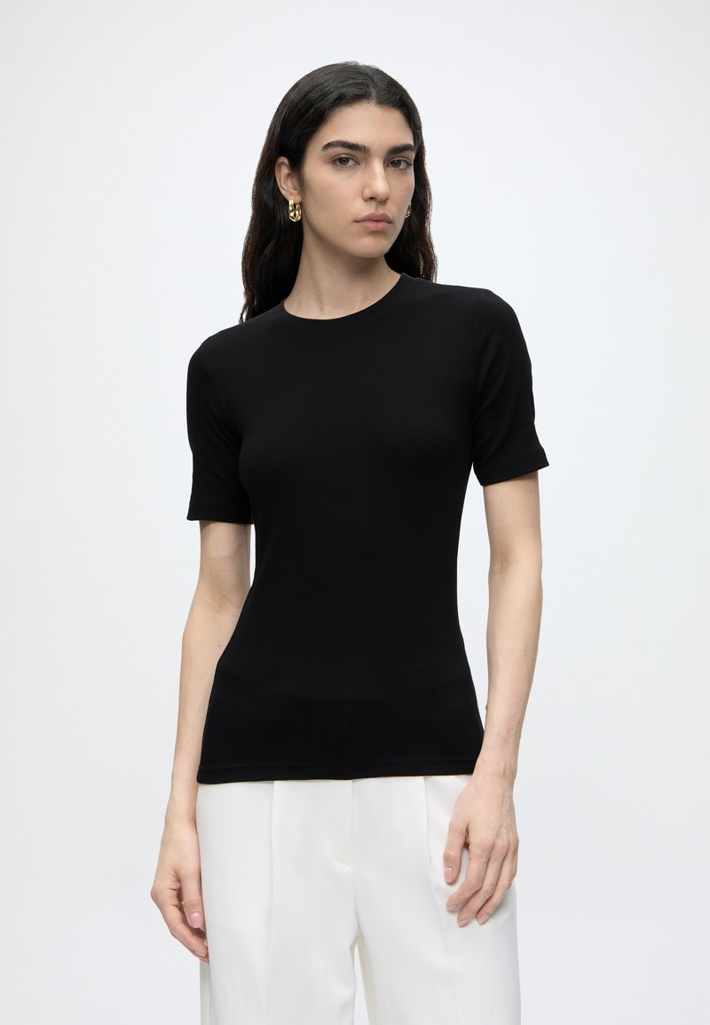 Γυναίκα με μακριά σκούρα μαλλιά που φοράει εφαρμοστό μαύρο κοντομάνικο τοπ και άσπρο παντελόνι σε απλό φωτεινό φόντο.