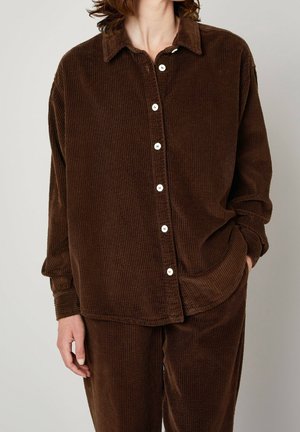 Femme portant une chemise boutonnée ample en velours côtelé marron et un pantalon assorti, une main dans la poche, debout devant un fond uni.