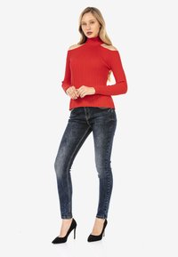 Roter gerippter Pullover mit kalten Schultern, kombiniert mit dunkelblauen Skinny Jeans und schwarzen High Heels. Hat einen hohen Kragen und ein strukturiertes Material.