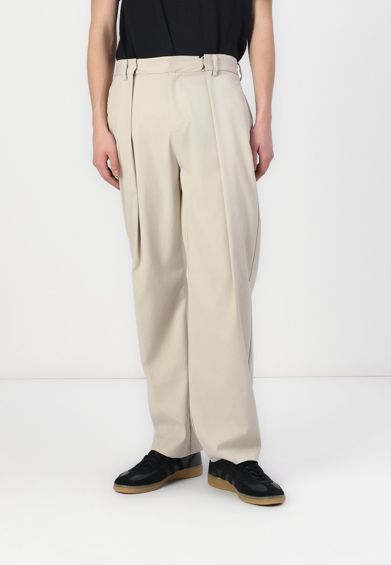 Beige Plissee-Hosen mit hoher Taille und Gürtelschlaufen, aus leichtem Stoff. Getragen mit schwarzen Sneakers mit einer Gummisohle.