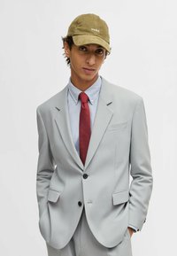 Light gray suit with tailored fit, paired with a blue dress shirt, dotted red tie, and olive green cap featuring white logo embroidery.