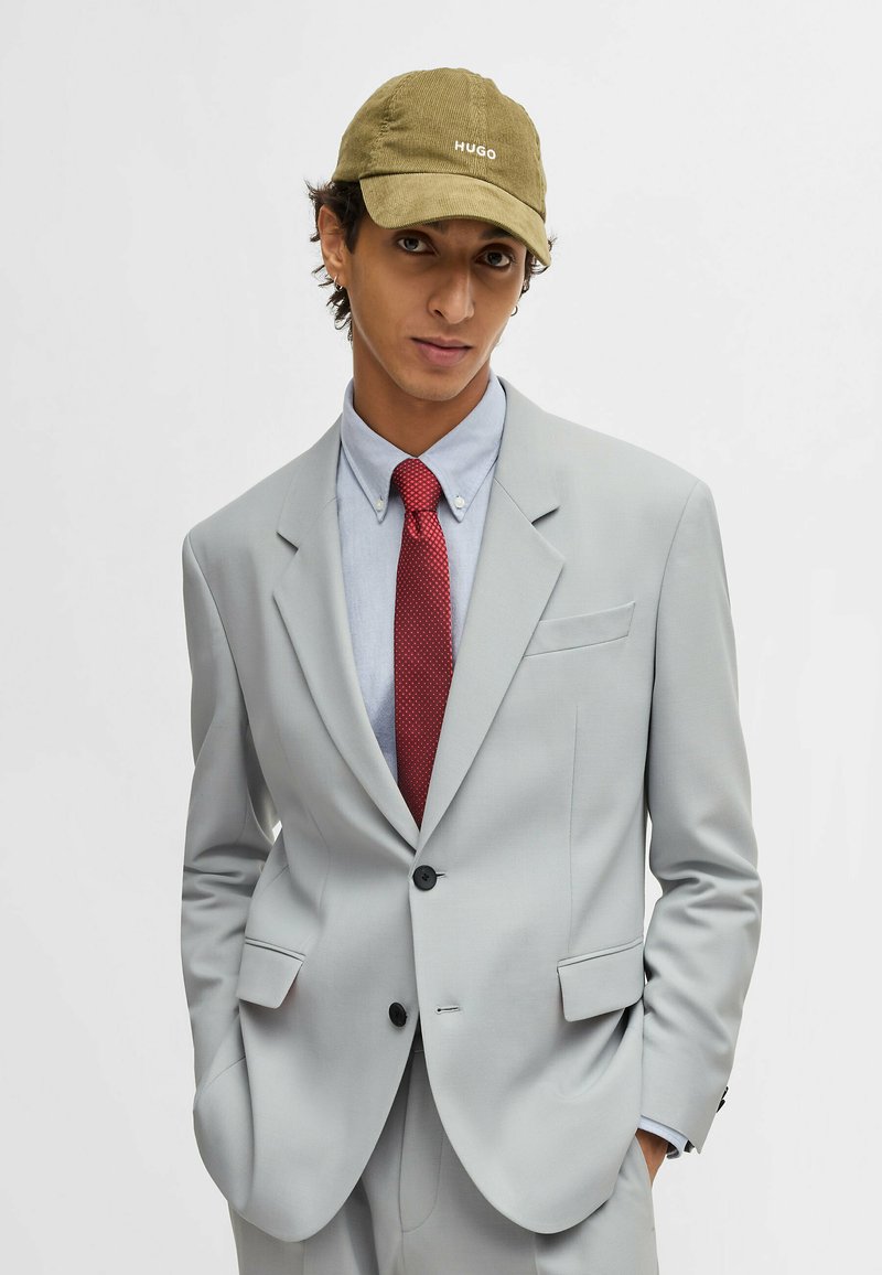 Light gray suit with tailored fit, paired with a blue dress shirt, dotted red tie, and olive green cap featuring white logo embroidery.