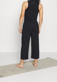 Haut noir sans manches et pantalon noir court en textile doux. Le pantalon est ample et possède des poches latérales. Pieds nus sur un tapis de yoga.