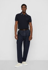 Polo di colore navy con colletto bianco, abbinato a pantaloni in denim scuro e sneakers di colore chiaro, caratterizzato da una vestibilità classica e una consistenza resistente.