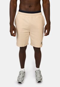 Beige katoenen shorts in een blend met een elastische tailleband, voorzien van een zijlogo-detail, vallend tot halverwege het dijbeen, in combinatie met grijze sportschoenen.