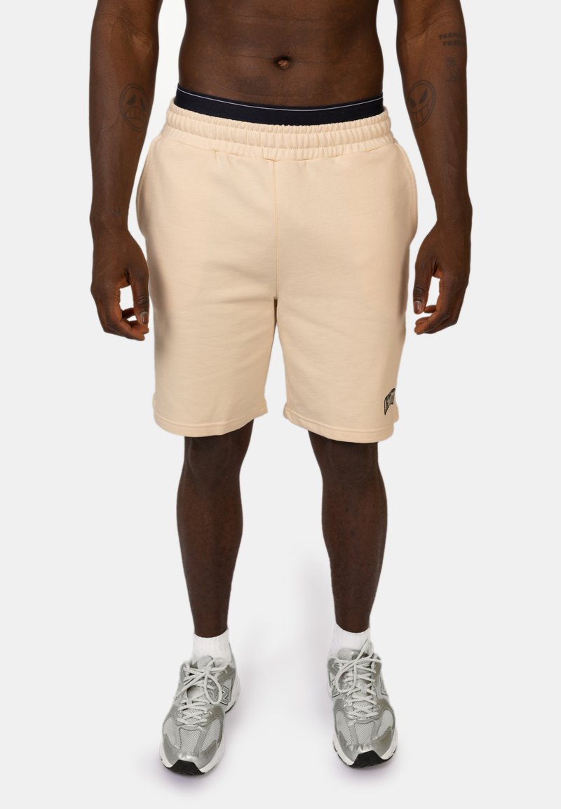 Beige katoenen shorts in een blend met een elastische tailleband, voorzien van een zijlogo-detail, vallend tot halverwege het dijbeen, in combinatie met grijze sportschoenen.