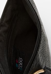 Musta teksturoitu clutch-laukku vetoketjukiinnityksellä, jossa on sileä kangasvuori ja sisätasku. Kultasävyinen metalliosat ja brändilogo näkyvät.