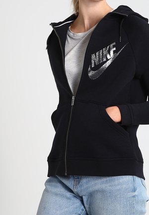 Femme portant un sweat à capuche noir Nike zippé avec un logo scintillant, un t-shirt gris clair en dessous et un jean bleu clair, les mains dans les poches.