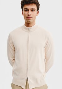 Jasnobeżowy sweter zapinany na zamek z wysokim kołnierzem, wykonany z miękkiej dzianiny, z długими rękawami i prostym dołem.