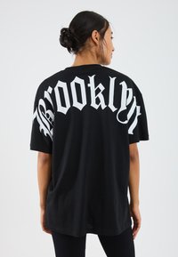 Svart oversized t-shirt med stor vit text "Brooklyn" på ryggen. Mjuk bomullsmaterial med avslappnad passform och korta ärmar.