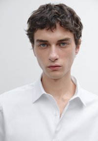 Weißes Hemd mit Knopfleiste aus Baumwolle, mit spitzem Kragen und glatter Textur, getragen von einem männlichen Model mit kurzen, lockigen Haaren.