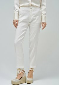 Pantalon en lin blanc à coupe slim, avec des poignets retroussés et des accents noués. Associé à des espadrilles compensées en beige clair ornées de broderies.