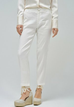 Pantalon en lin blanc à coupe slim, avec des poignets retroussés et des accents noués. Associé à des espadrilles compensées en beige clair ornées de broderies.