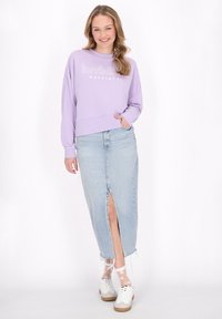 Crop top viola chiaro con il testo "felicità invisibile", abbinato a una gonna di jeans chiara con uno spacco frontale e orlo sfrangiato. Sneakers bianche.