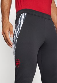 Pantalones deportivos negros con una textura suave, que presentan un patrón de zigzag blanco en el costado y un logo rojo de Arsenal en la parte inferior de la pierna.