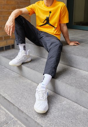 Personne en T-shirt orange avec logo noir, pantalon noir, chaussettes blanches et baskets blanches assise sur des marches en béton, un bras reposant derrière.