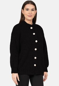 Młoda kobieta o prostych brązowych włosach nosi czarny sweter z dzianiny z dużymi srebrnymi guzikami, stoi na tle jednolitego białego tła.