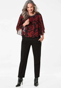 Blusa nera a fiori con maniche fluenti e motivi rossi, abbinata a pantaloni neri su misura e decolleté neri a punta. Tessuto liscio e leggero.