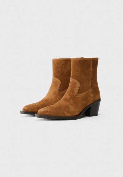 Cizme de gleznă din suede maro, cu vârf ascuțit, toc înclinat și accente de cusătură. Textură netedă, cu o potrivire structurată și un design subtil.