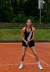 Teniso žaidėja juodais sportiniais drabužiais, laikanti raketę ant molio aikštelės. Ji turi laisvus, garbanotus plaukus ir baltus sportinius batus. Fone matyti žali medžiai.