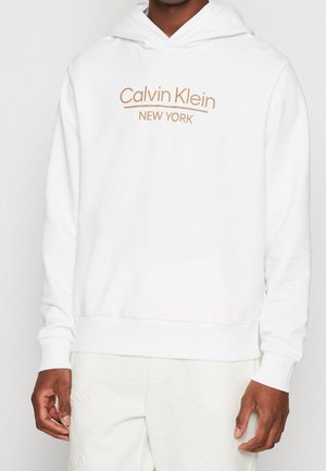 Mann trägt weißen Calvin Klein Hoodie mit dem beigen Schriftzug "Calvin Klein New York" auf der Vorderseite, kombiniert mit weißen strukturierten Hosen.