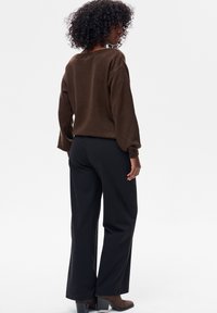 Kobieta o kręconych włosach, ubrana w brązowy sweter, czarne spodnie o szerokich nogawkach oraz brązowe buty na obcasie, stojąca na białym tle.