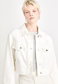 White cropped denim jacket with long sleeves, button front, chest pockets, and silver-tone hardware. Worn over a grey top. Smooth texture.