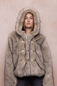 Woman wearing a beige fur hooded coat with metal clasps, standing against a plain light pink background.
