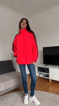 Raudonas Adidas vėjuotas striukė su juodomis juostomis, pilnas užtrauktukas, aukštas apykaklė; dėvima ant mėlynų siaurų džinsų ir baltų sportbačių.