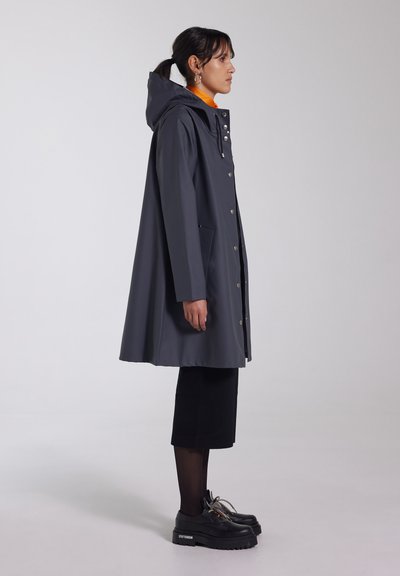 Grijze regenjas met capuchon, drukknoopsluitingen en voorzakken. Gemaakt over een oranje top en gecombineerd met een zwarte rok en dikke plateau schoenen.