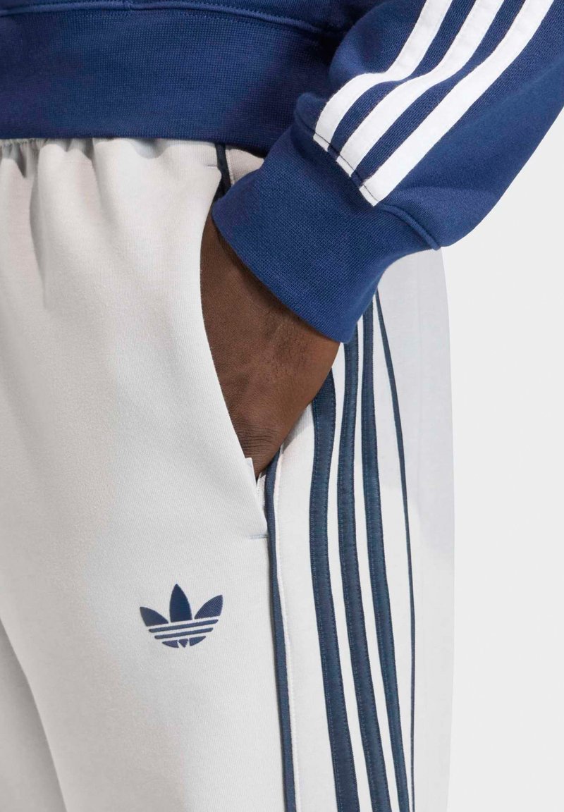 Main à la peau foncée dans la poche d’un pantalon blanc Adidas avec des rayures bleu marine, associée à une manche bleu marine avec des rayures blanches.