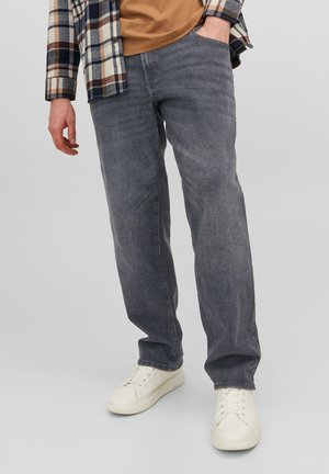 Homme portant un jean droit gris, des baskets blanches, une chemise marron, et une veste à carreaux bleue, blanche et marron, debout avec une main dans la poche.