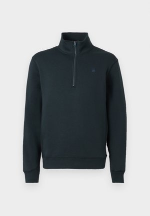 Sweat-shirt zippé vert foncé en tissu doux, avec un col montant, des poignets côtelés et un petit logo sur la poitrine.