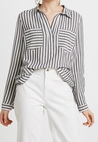 Camicia a maniche lunghe a righe grigie e bianche, con collo, due tasche frontali e vestibilità ampia, abbinata a pantaloni bianchi.