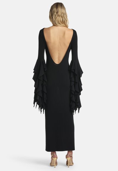 Mujer vistiendo un largo vestido negro con escote en V profundo en la espalda y mangas acampanadas con volantes en capas, combinado con sandalias doradas de tacón alto, de espaldas.