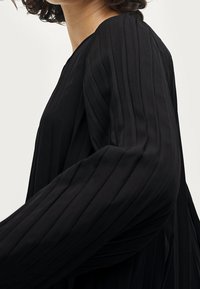 Musta laskostettu mekko, jossa on pitkät hihat. Kankaassa on sileä pinta, pystysuorat raidat ja pyöreä pääntie.