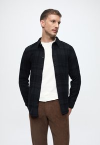 Μαύρο καρό πουκάμισο με κουμπιά από μαλακό υλικό, φορεμένο πάνω από ένα λευκό μπλουζάκι με στρογγυλή λαιμόκοψη, συνδυασμένο με καφέ παντελόνι καμισού.