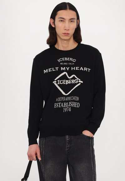 Pull noir avec texte brodé blanc : "LA GLACE FOND MON COEUR" et logo. Manches longues, col rond et ourlet côtelé. Porté avec un jean gris.