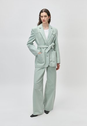 Γυναίκα με μακριά σκουρόχρωμα μαλλιά που φοράει σακάκι μέντας με ζώνη και ταιριαστό φαρδύ παντελόνι πάνω από λευκό τοπ, στέκεται μπροστά σε απλό ανοιχτό φόντο.