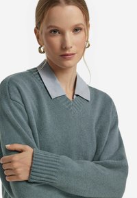 Himmelblaues V-Ausschnitt-Pullover mit gerippten Bündchen und Saum, über einem himmelblauen Hemd mit Kragen getragen. Glatte Textur, lässige Passform.