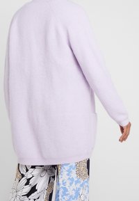 Liliowy sweter kardigan o luźnym kroju z bocznymi kieszeniami; charakteryzuje się teksturowanym materiałem oraz długimi rękawami. Tył ma prosty fason.