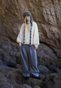 Krémový, nadrozmerný sveter s balónovými rukávmi, kombinovaný s voľnými sivými nohavicami a klobúkom s kožúškom a modrými akcentmi.