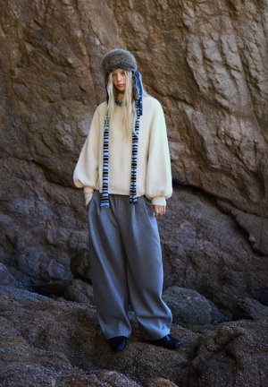 Kremowy, oversize'owy sweter z balonowymi rękawami, zestawiony z luźnymi szarymi spodniami i czapką z futrzanym wykończeniem w niebieskie akcenty.