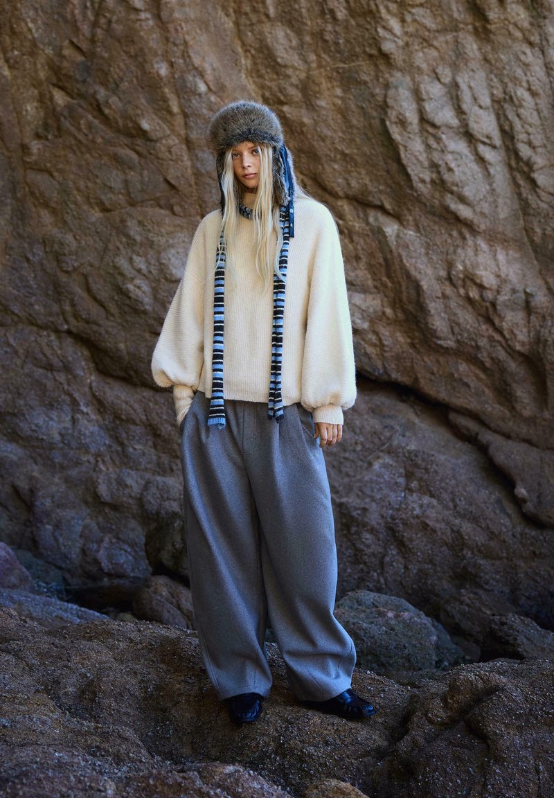 Krémový, nadrozmerný sveter s balónovými rukávmi, kombinovaný s voľnými sivými nohavicami a klobúkom s kožúškom a modrými akcentmi.