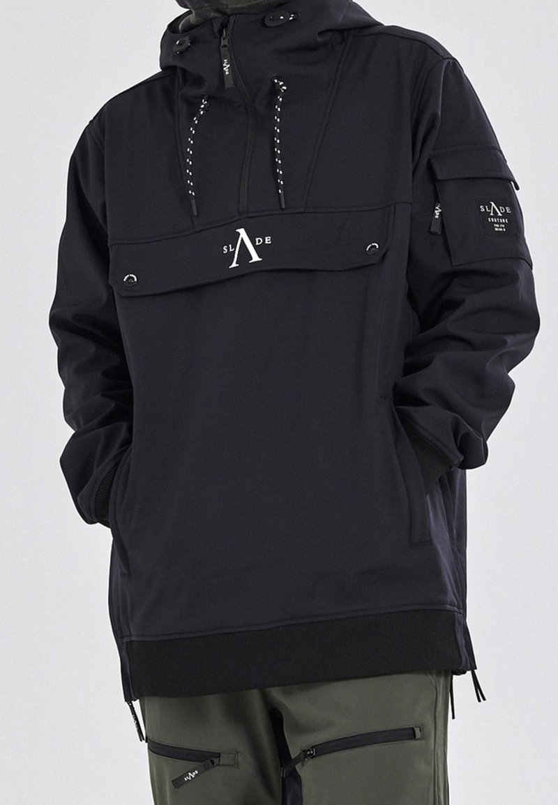 Personne portant une veste à capuche noire avec le logo SLΛDE sur le devant et la poche de manche, associée à un pantalon vert avec des poches zippées. Mains dans les poches.